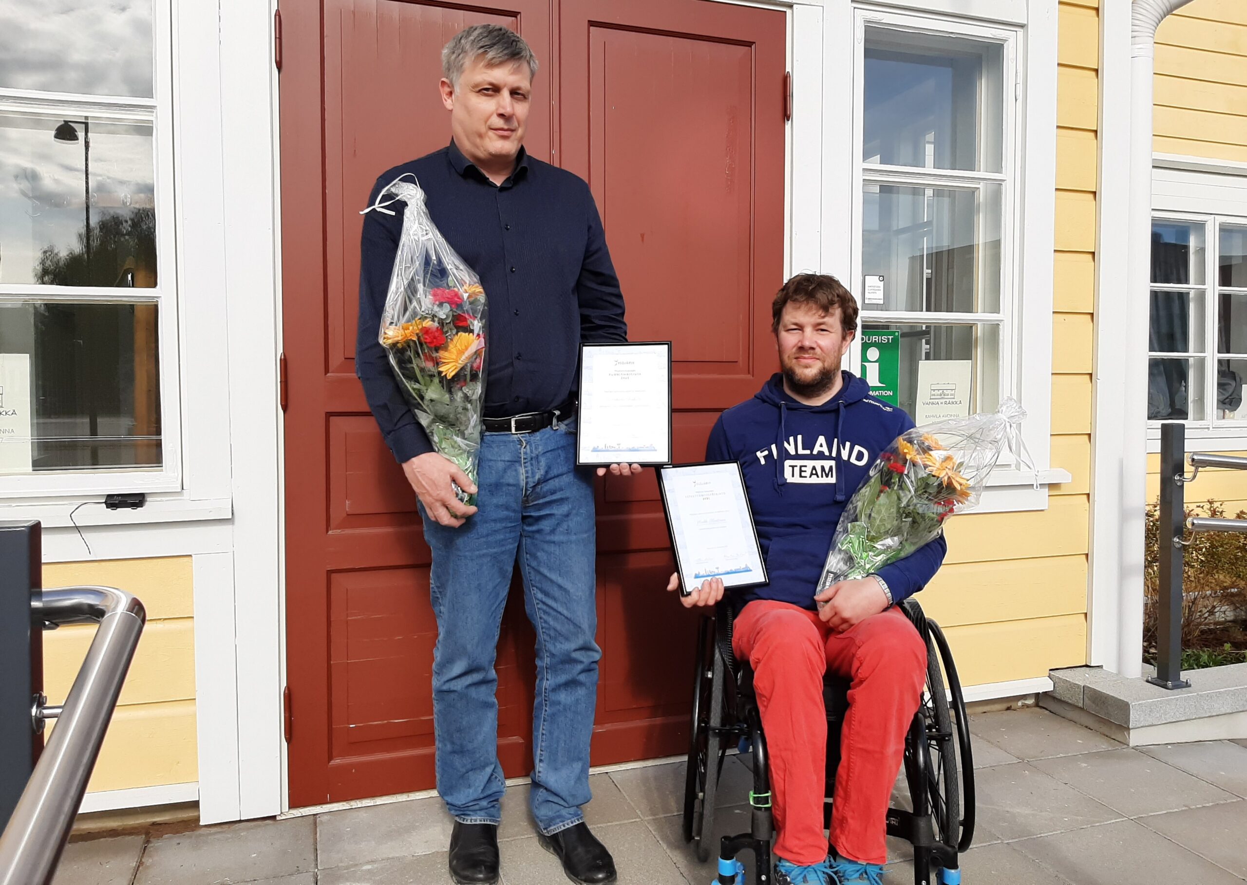 Kaksi miestä talon edustalla kukkakimput ja kunniakirjat käsissään