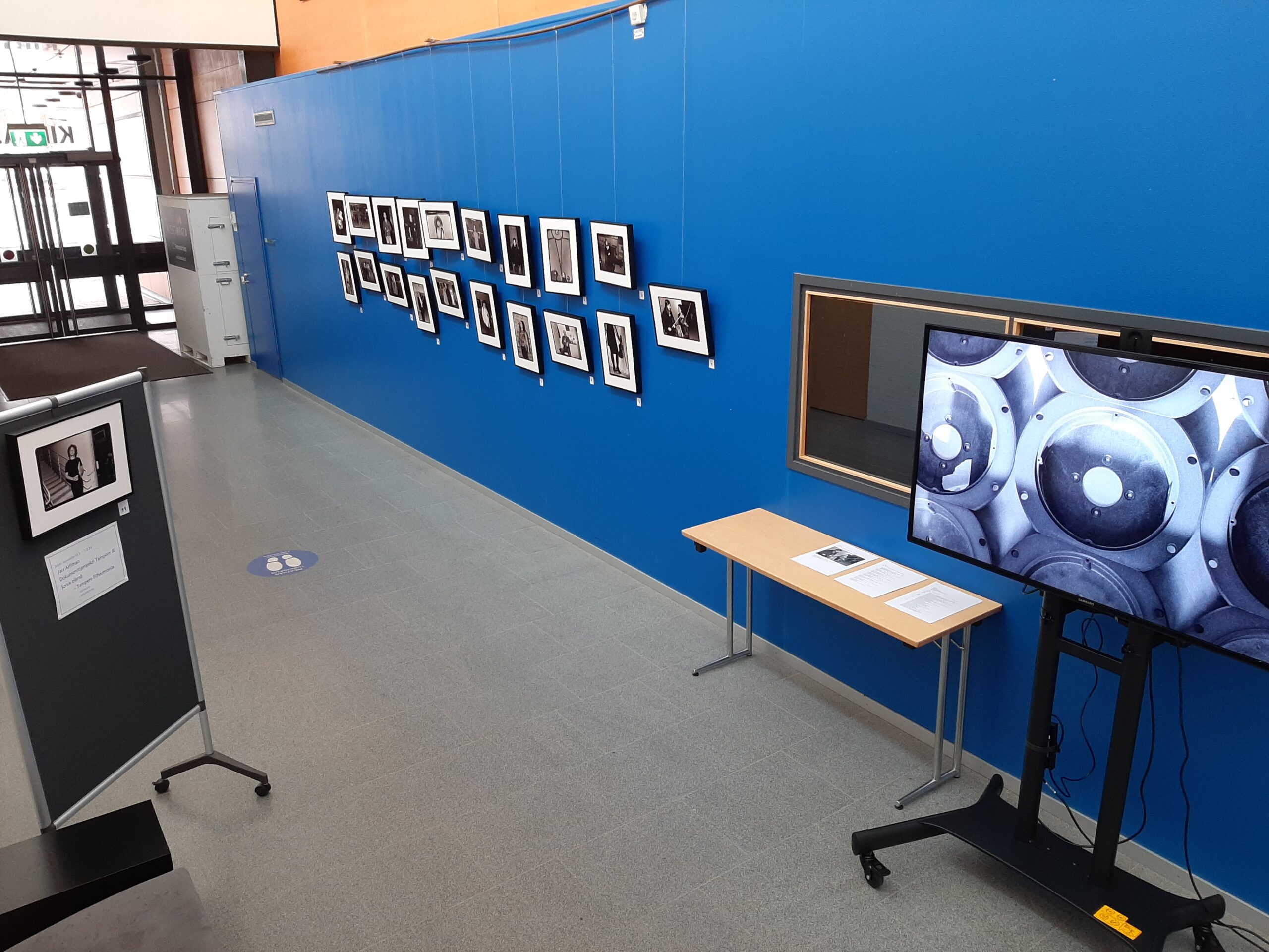Kuva näyttelystä, kehystettyjä valokuvia seinällä sekä näyttö lyhytelokuvan katselua varten.