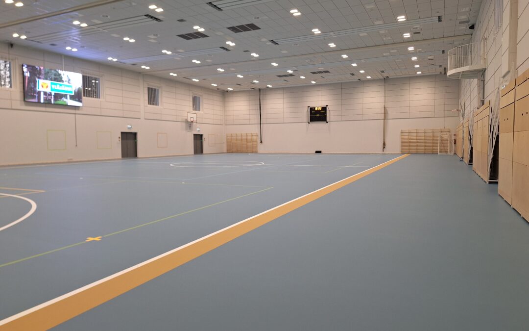 Kuvassa Siltatien liikuntahallin palloilusali, jonka sinisellä lattipohjalla paljon viivoituksia. Katossa valoja ja seinällä LED-näyttö.