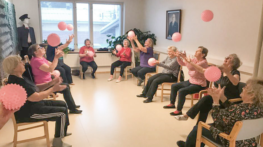 Ryhmä tekemässä yhdessä tuolijumppaa istuen ja heittäen samalla ilmaan vaaleanpunaisia palloja.