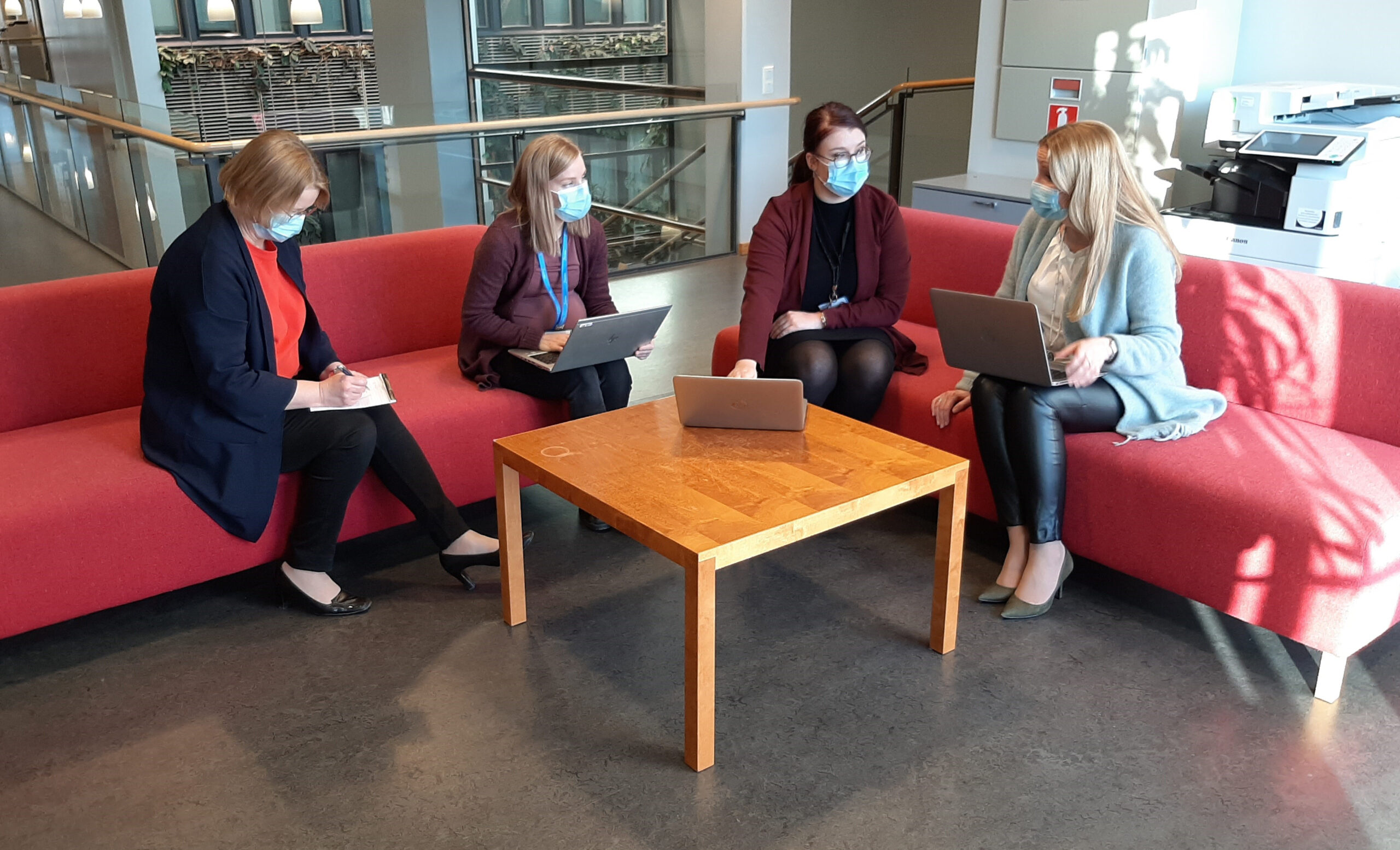 Neljä naista istuu aulatilan neuvottelunurkkauksessa kannettavien tietokoneiden kanssa