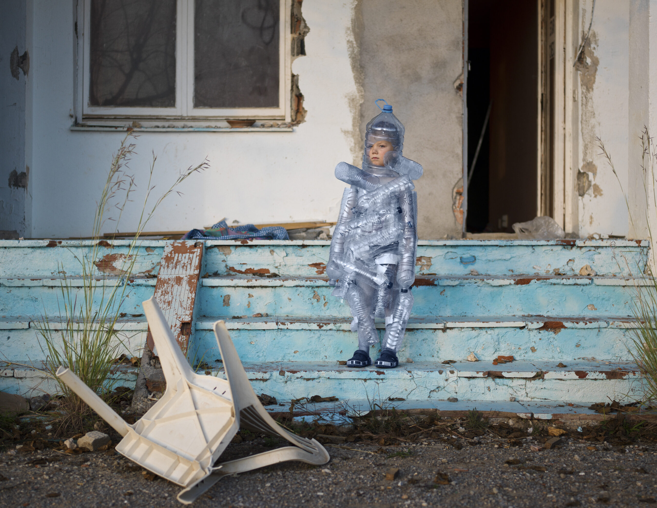 Valokuvateoksessa lapsi seisoo ränsistyneen talon rappusilla muovipulloihin pukeutuneena.
