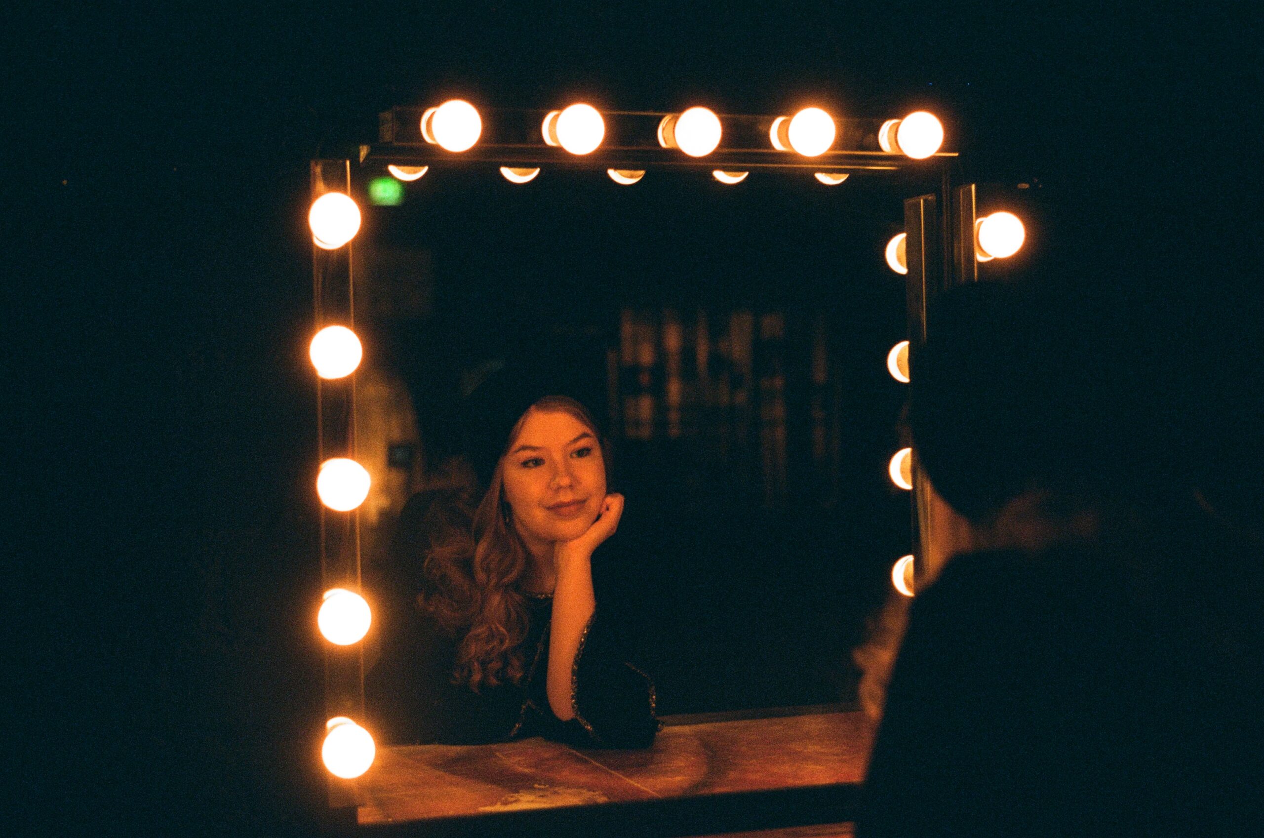 Nainen katsoo peiliin, jota ympäröivät valot. Teatterin takahuoneen meikkauspeili.