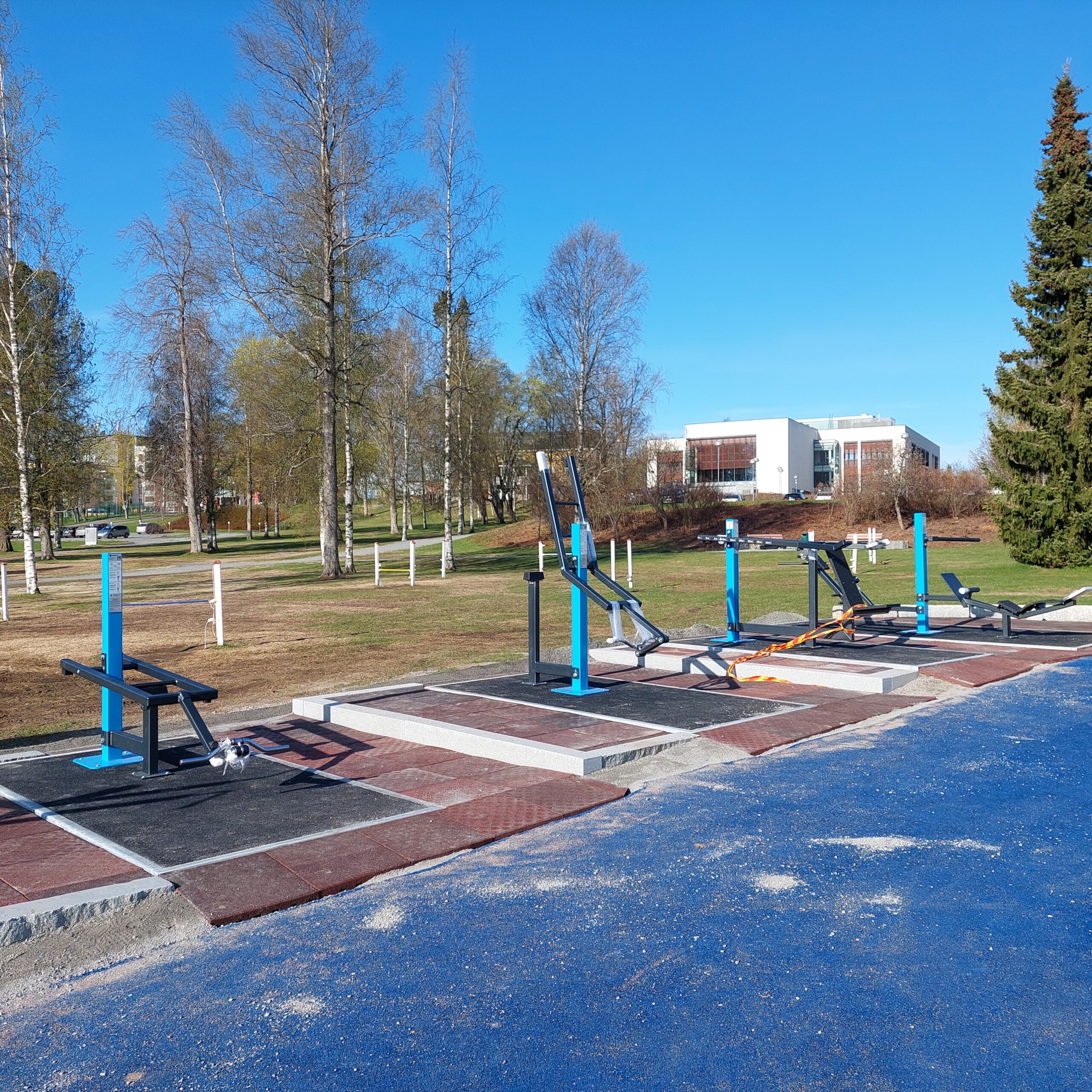 Neljä sinistä ulkokuntosalilaitetta puistossa, taustalla Ylöjärven kaupungintalo.