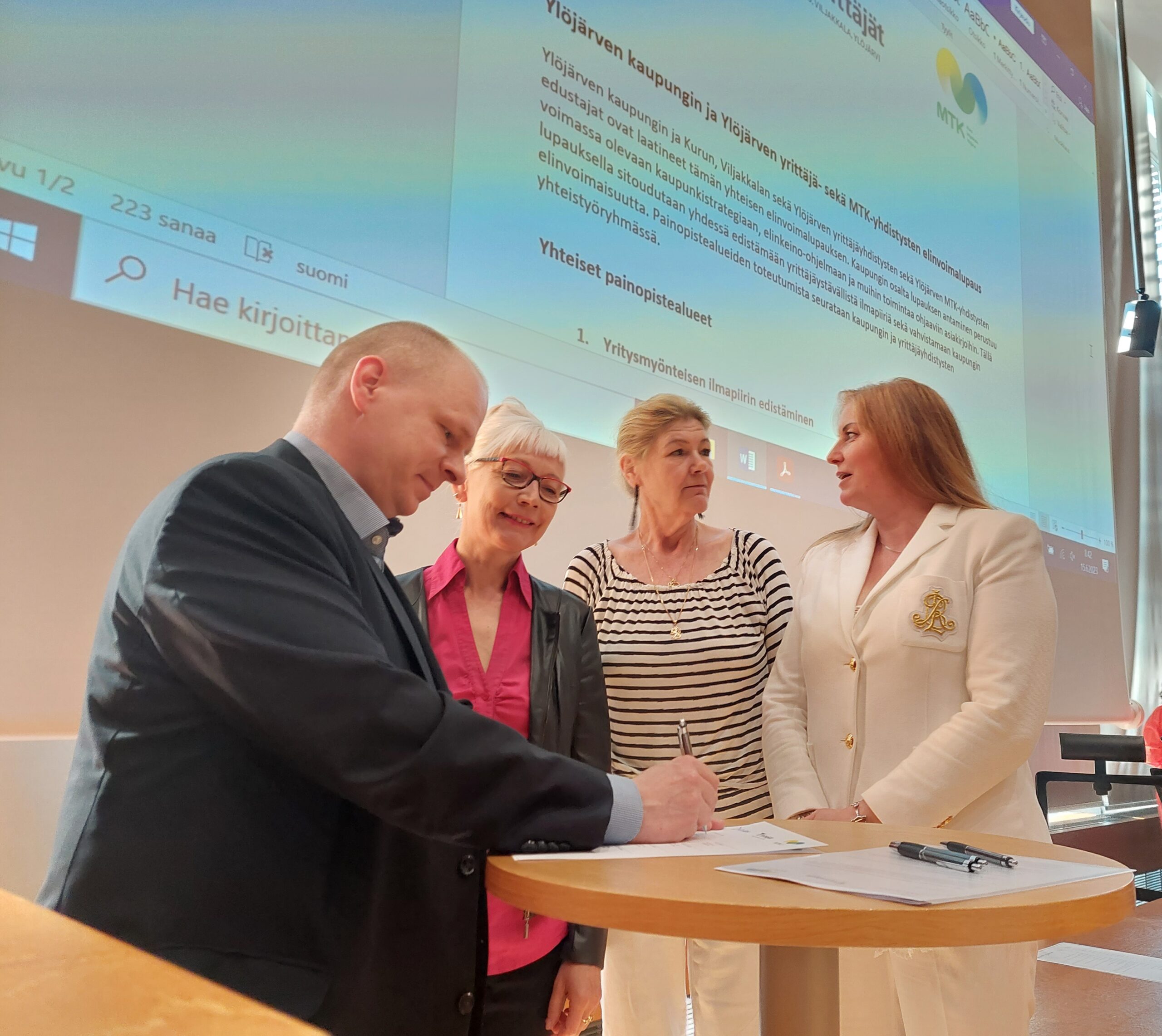 Kaupunginjohtaja allekirjoittaa sopimuksen, kolme naista seisoo vieressä.